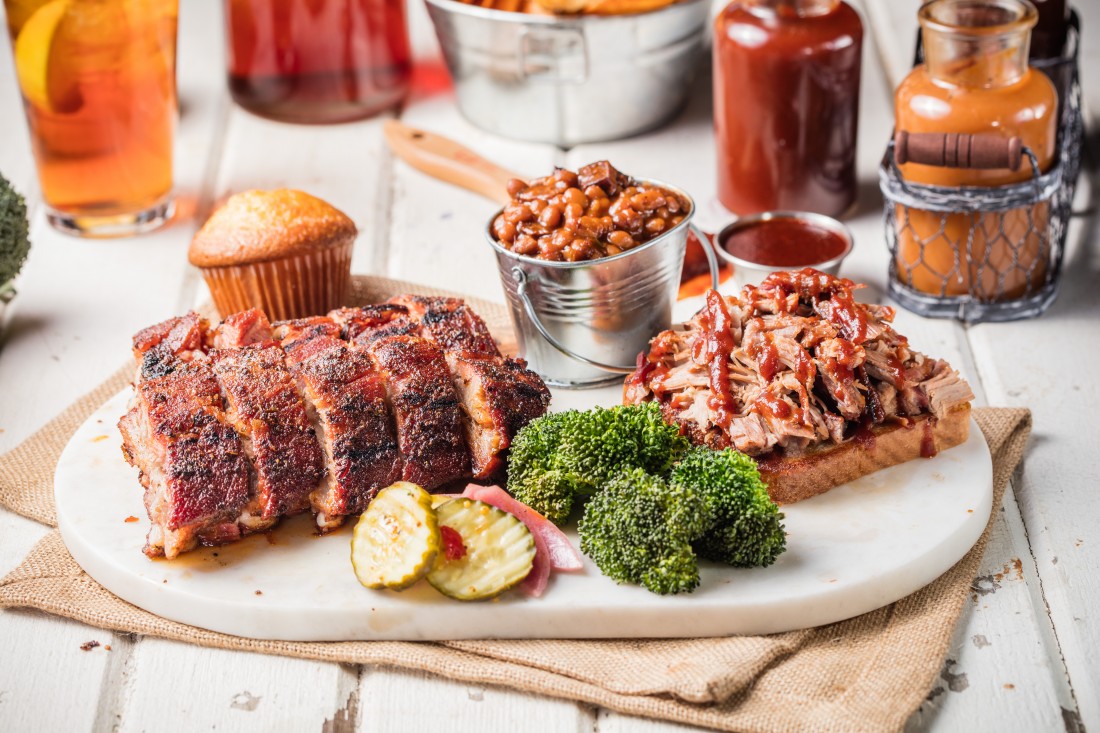 A Famous Dave's Two-Meat Combo, served with a cornbread muffin, pickles, pickled onions, broccoli, barbecue sauce, and baked beans in a small silver pail