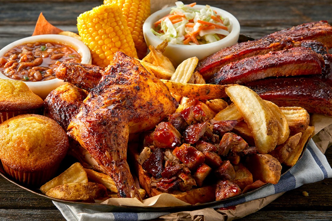 Image of Famous Dave's All-American BBQ Feast® with chicken legs, ribs, corn on the cob, cornbread, wedge fries, baked beans, and cole slaw all stacked on top of each other