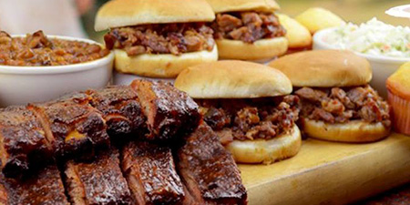 In the foreground sits sliced BBQ meats and two chopped pork sandwiches in burger buns, behind them sits a bowl of baked beans and two more chopped pork sandwiches