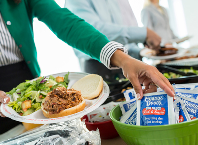 Woman with a plate of food reaching over to grab a packet of Famous Dave's BBQ sauce